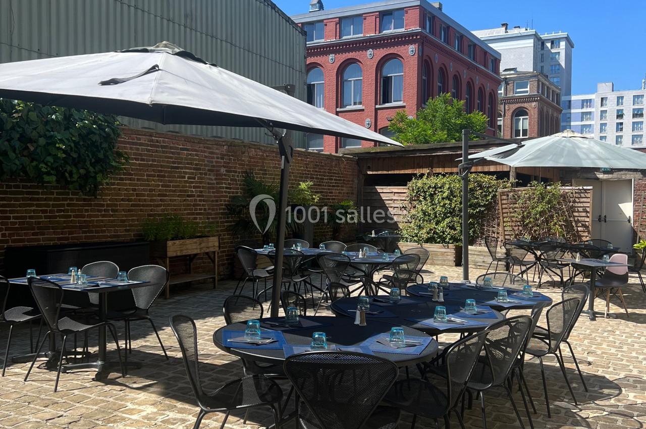 Terrasse extérieure avec tables et chaises en métal, parasols ouverts, entourée de murs en briques et bâtiments urbains.