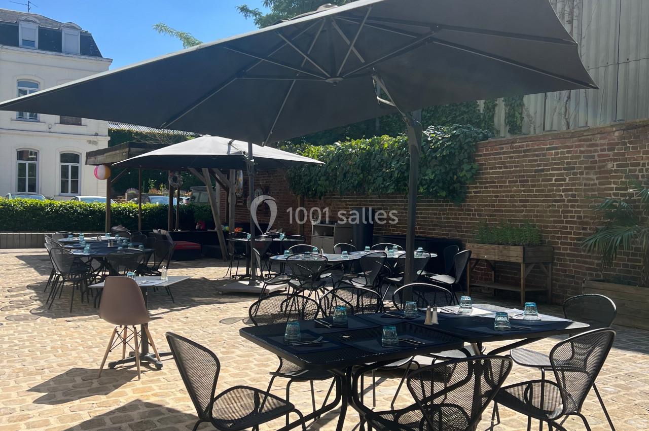 Terrasse extérieure ensoleillée avec tables et chaises noires, parasols ouverts et murs en briques en arrière-plan.