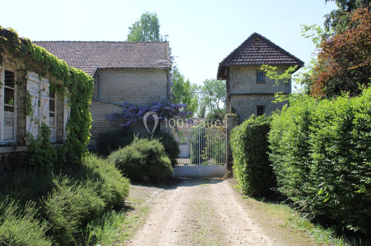Chemin de terre bordé de verdure menant à une maison en pierre avec portail et végétation fleurie.