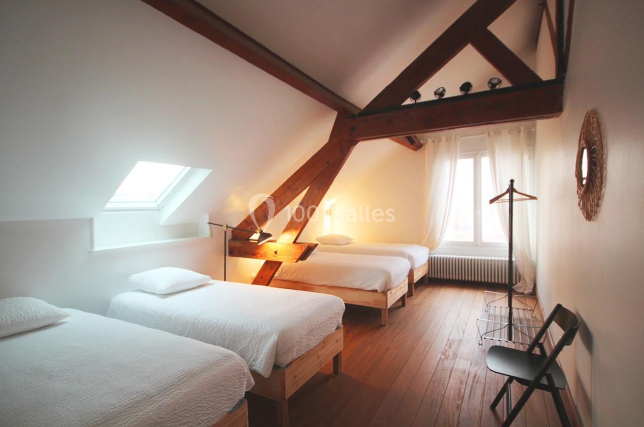 Chambre mansardée avec trois lits simples, poutres apparentes, lumière naturelle et mobilier minimaliste.