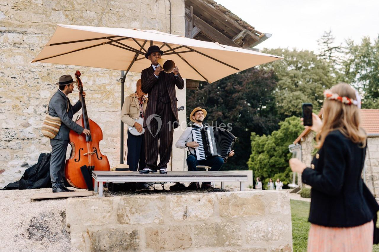 Un groupe de musiciens joue en plein air sous un parasol, avec une femme prenant une photo au premier plan.