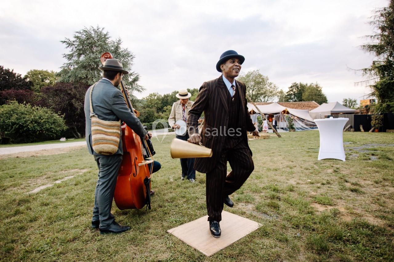 Un homme en costume danse sur une planche en plein air, accompagné de musiciens jouant de la contrebasse.
