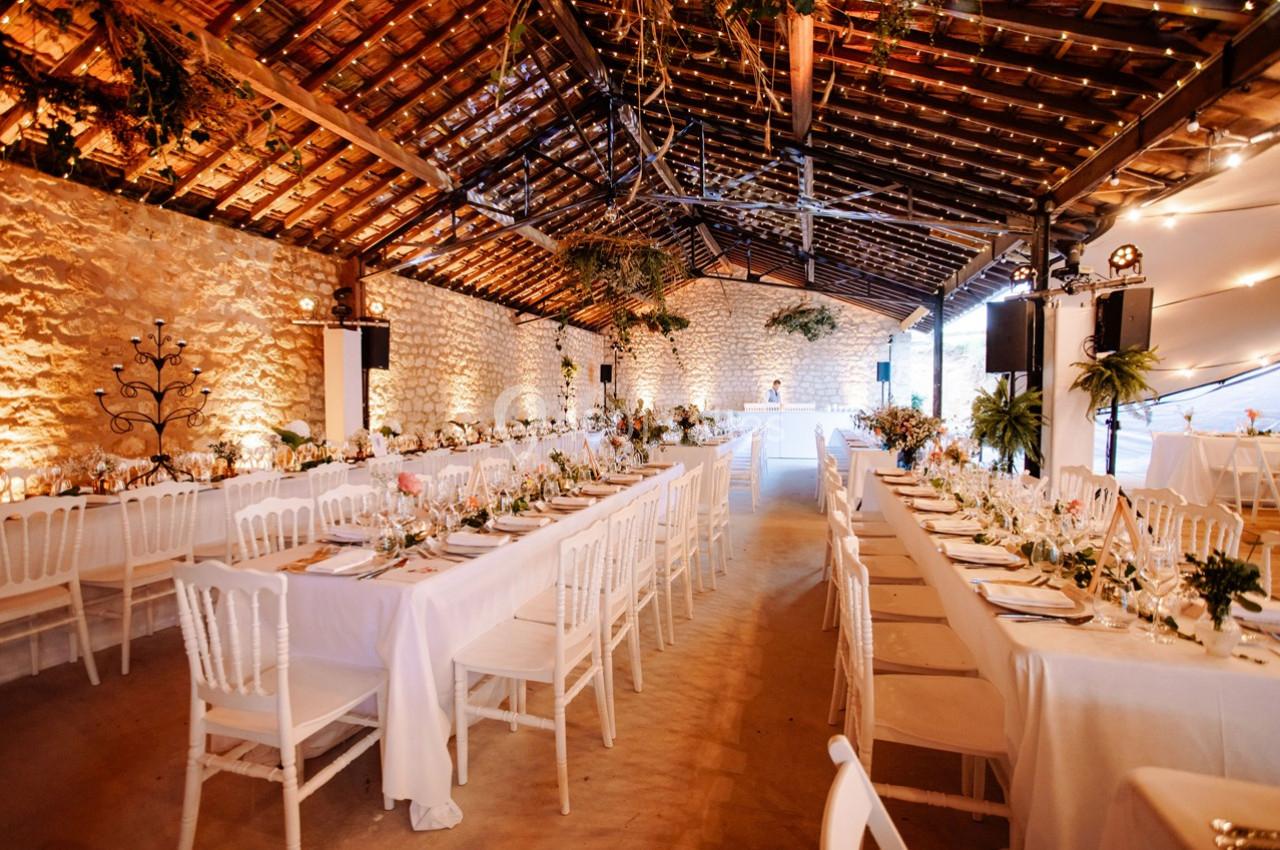 Salle de réception décorée avec des tables alignées, nappes blanches, chaises et guirlandes lumineuses suspendues.