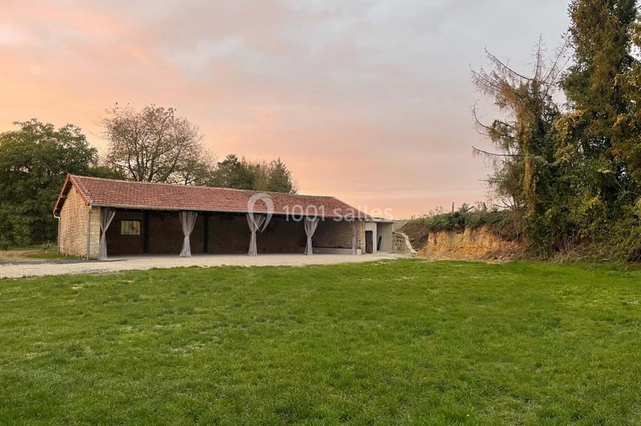 Bâtiment en pierre avec toit en tuiles, ouvertures en arcade, entouré de pelouse et d'arbres sous un ciel au coucher du…