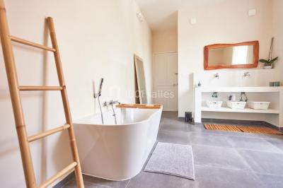 Salle de bain lumineuse avec baignoire moderne, échelle en bois, miroir incliné et double vasque sur meuble blanc.