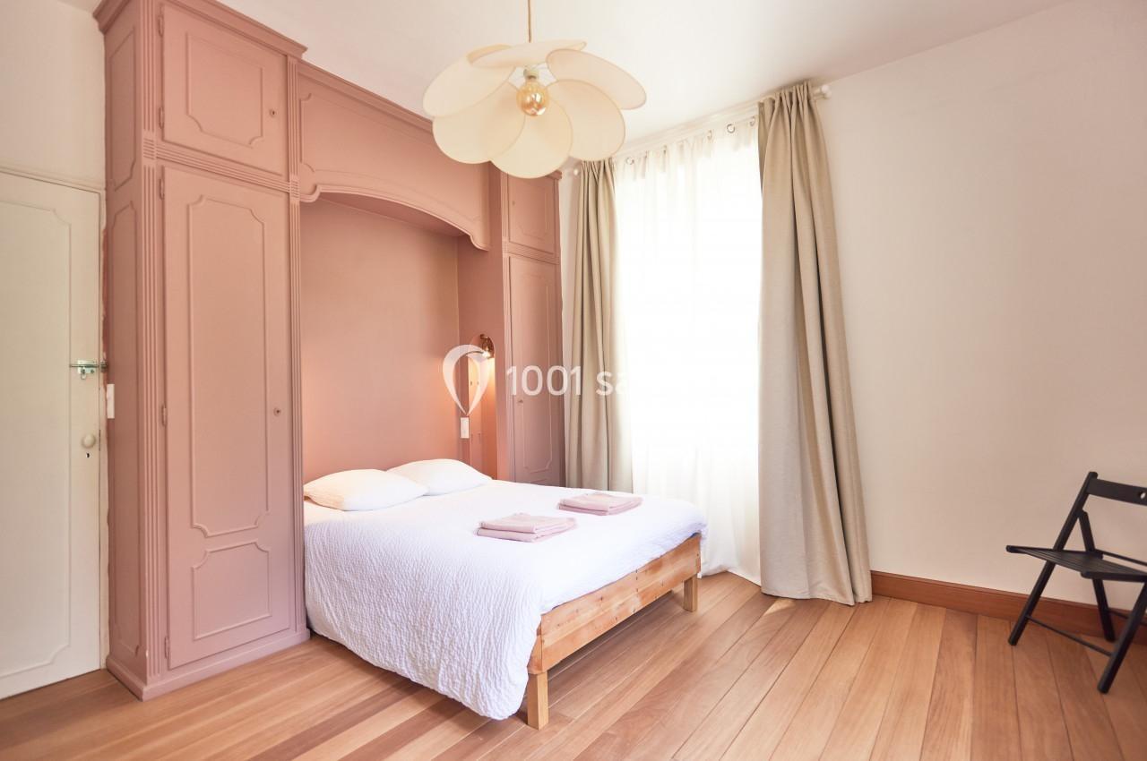 Chambre lumineuse avec lit double, armoire encastrée rose, parquet en bois et rideaux beiges près d'une fenêtre.
