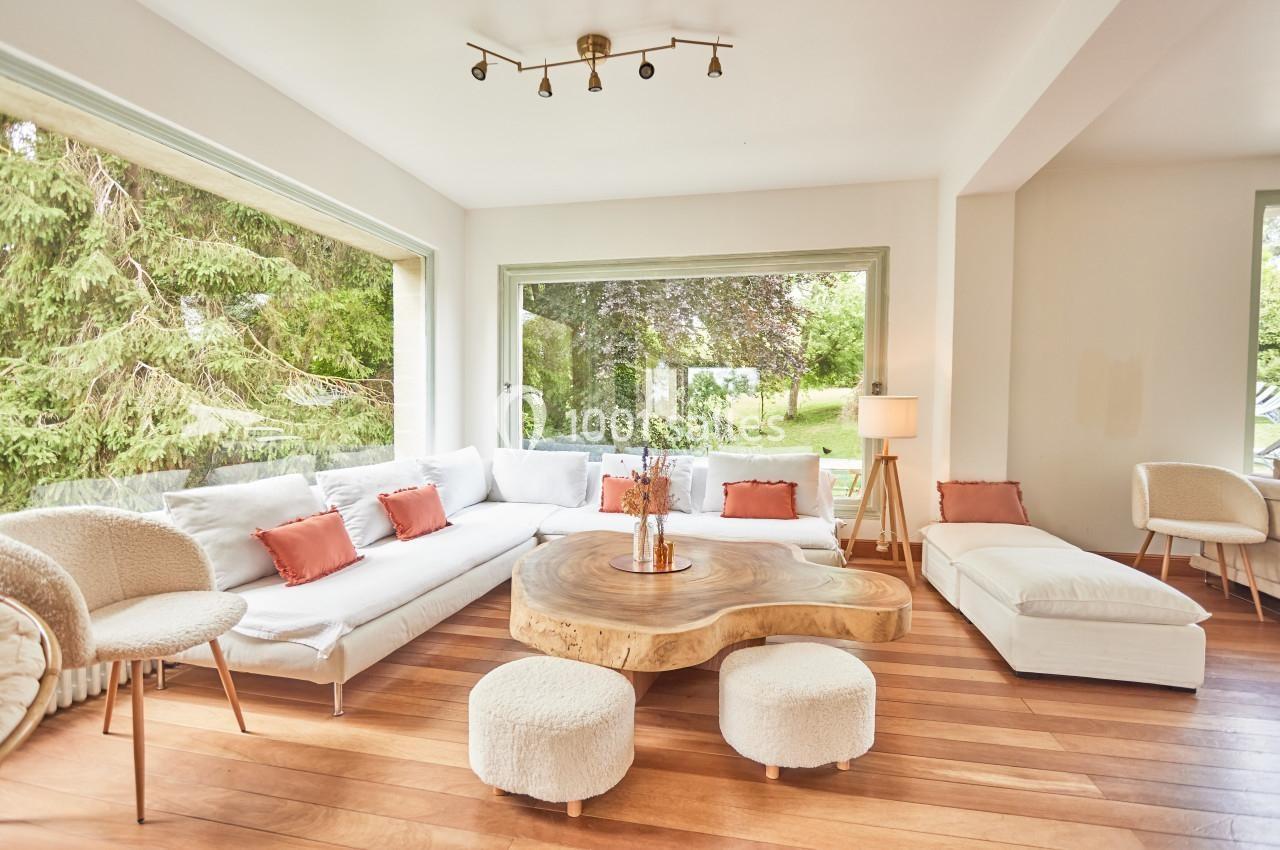 Salon lumineux avec canapé blanc, coussins orange, table basse en bois sculpté et grandes fenêtres donnant sur un jardin.