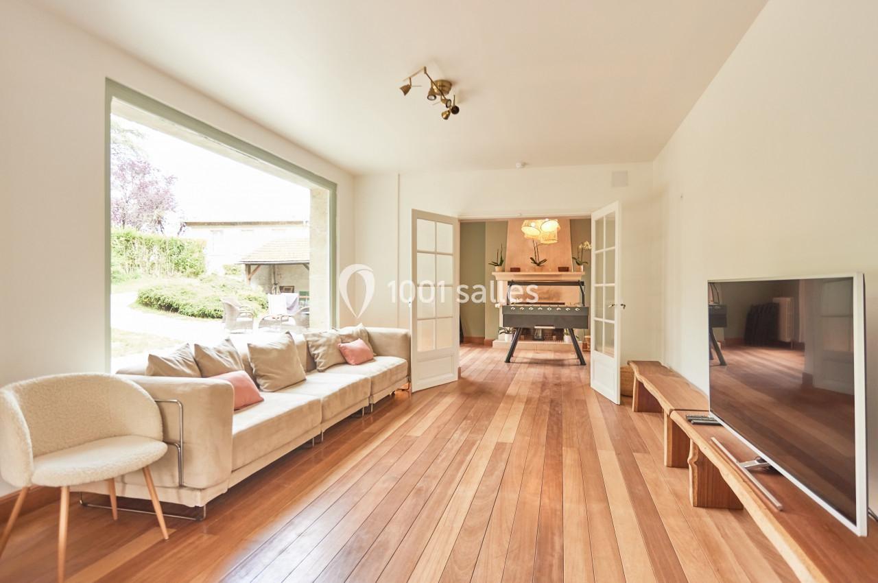 Salon lumineux avec parquet en bois, canapé beige, télévision et vue sur une salle à manger par des portes vitrées.
