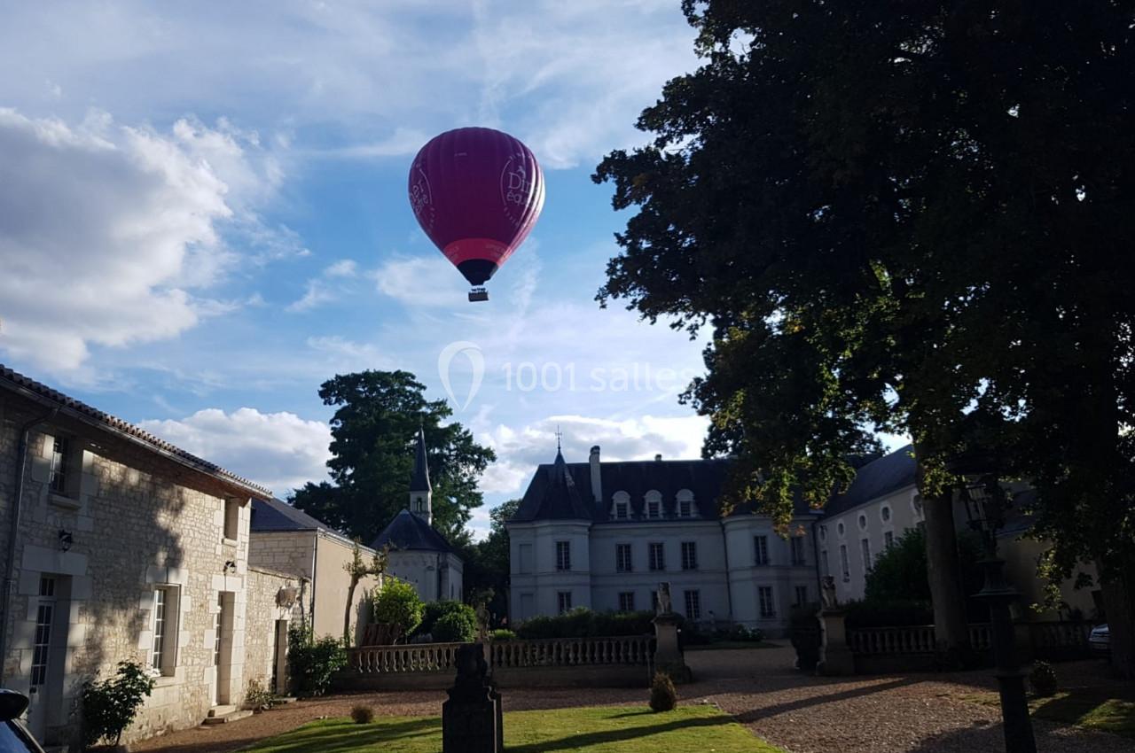Location salle Assay (Indre-et-Loire) - Chateau de Basché #7 Montgolfière rouge flottant au-dessus d'une cour avec bâtiments anciens et arbres sous un ciel partiellement nuageux.