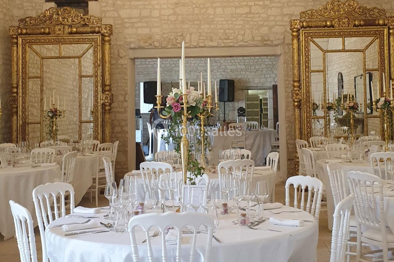 Location salle Assay (Indre-et-Loire) - Chateau de Basché #3 Salle de réception élégante avec tables rondes dressées, chaises blanches et grands miroirs dorés sur les murs en pierre.
