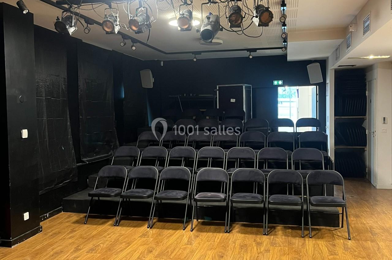 Salle de spectacle avec rangées de chaises noires vides, éclairage de scène suspendu et sol en parquet.
