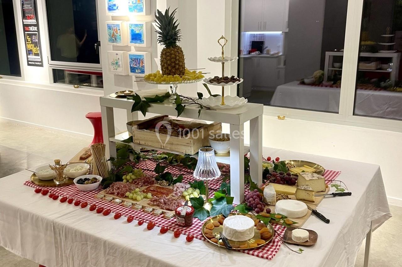 Table dressée avec des fromages, charcuteries, fruits et desserts, décorée de nappes à carreaux rouges et éléments végétaux.
