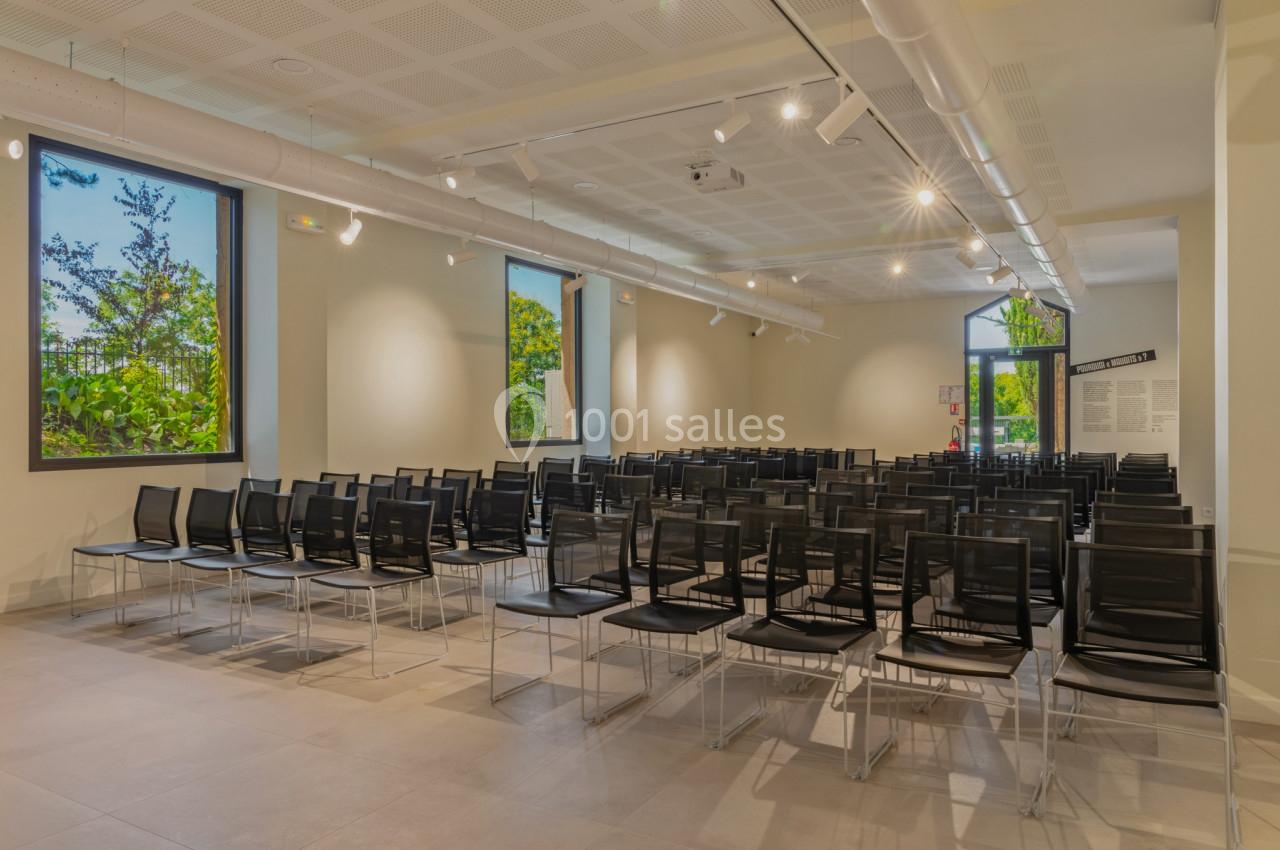 Salle lumineuse avec rangées de chaises noires alignées, grandes fenêtres laissant entrer la lumière naturelle.