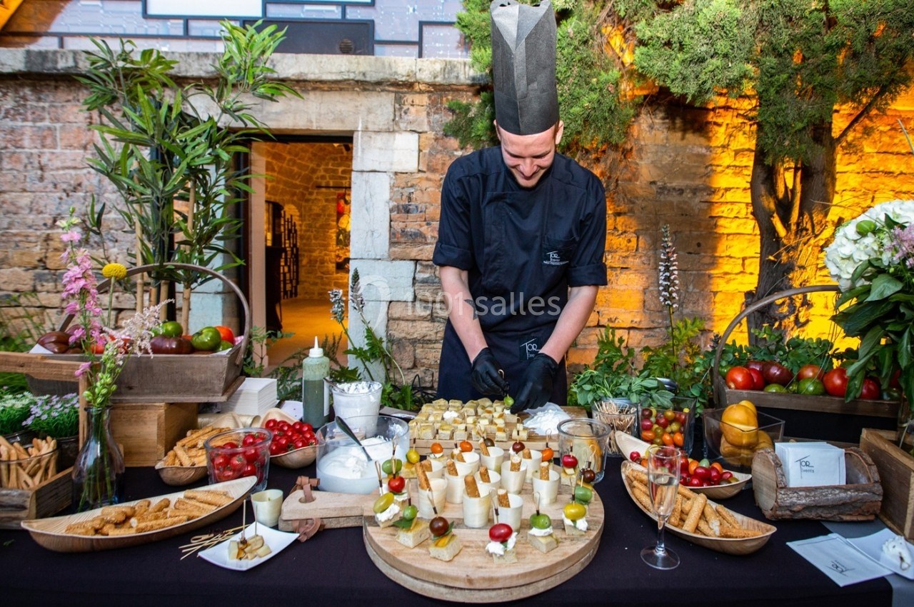 Un chef en tenue noire prépare un buffet de bouchées apéritives sur une table décorée en extérieur.