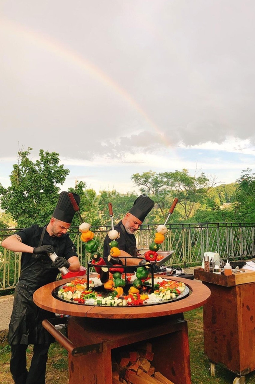 Deux chefs cuisinent des brochettes de légumes et de viande sur un barbecue circulaire en plein air sous un arc-en-ciel.