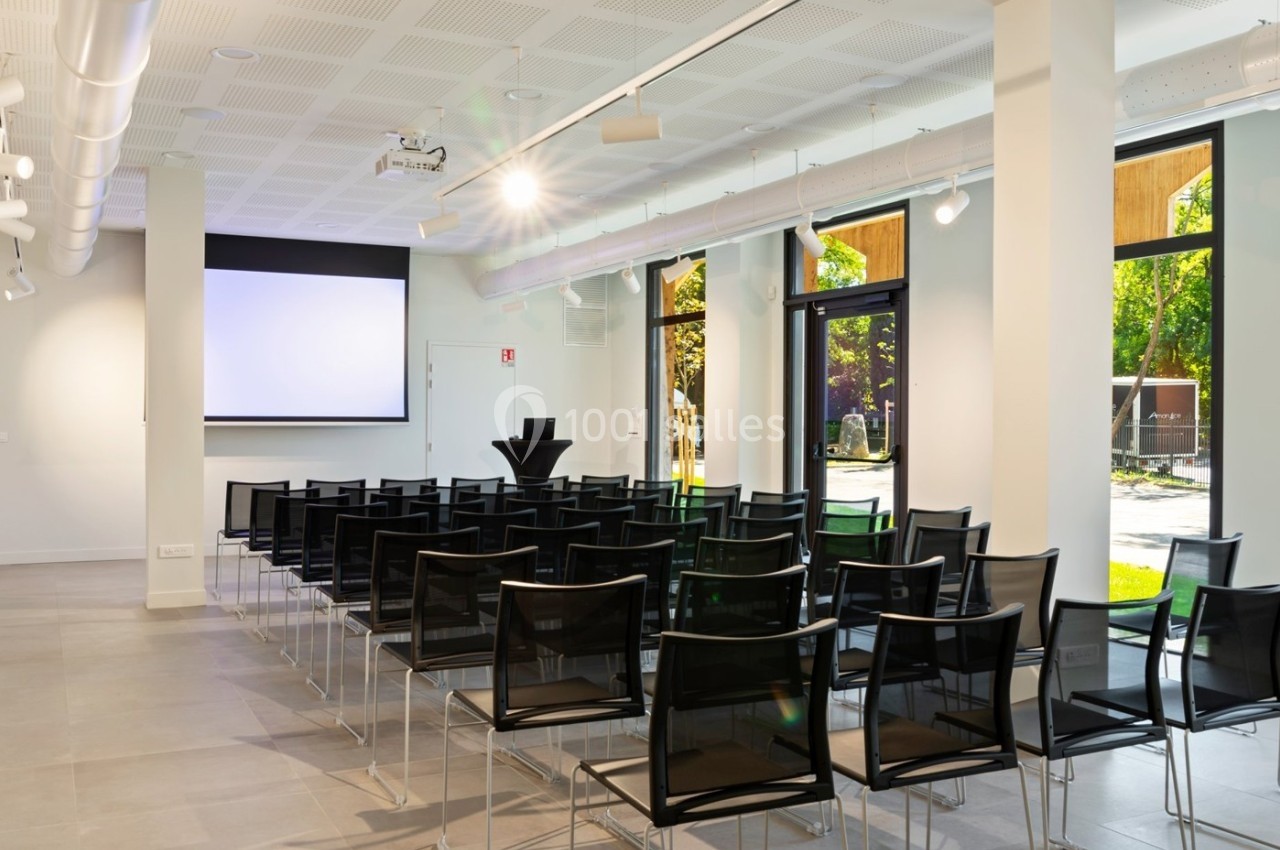 Salle de conférence lumineuse avec des chaises noires alignées, un écran de projection et de grandes fenêtres donnant sur l…