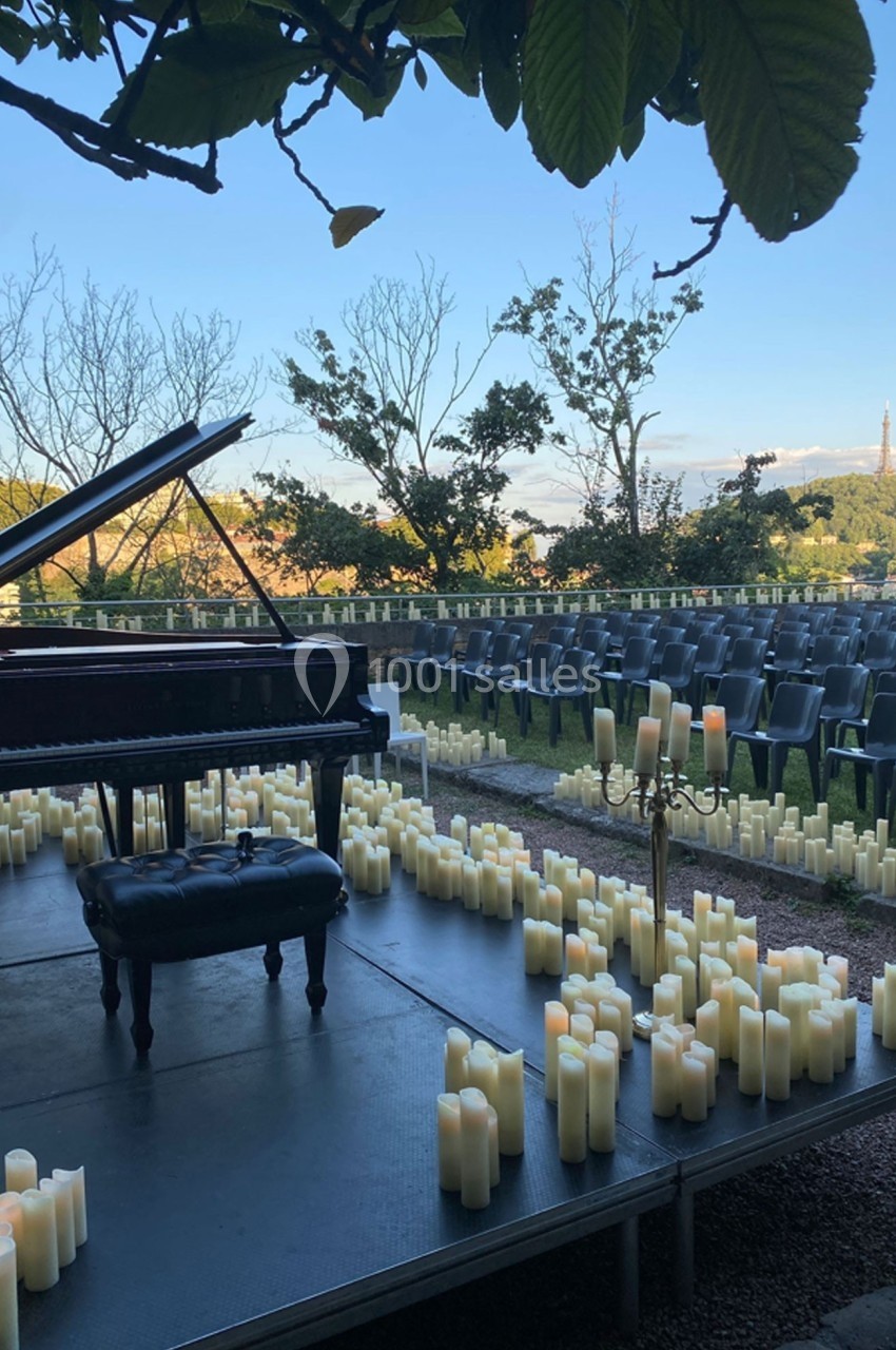 Scène extérieure avec un piano à queue entouré de bougies, des rangées de chaises vides en arrière-plan.