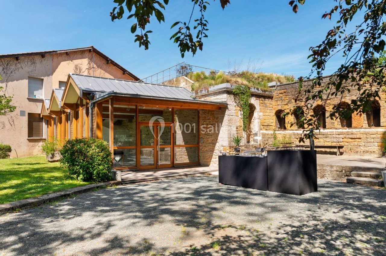 Entrée d'un bâtiment moderne avec façade en bois, entouré de verdure et de ruines en pierre sous un ciel dégagé.