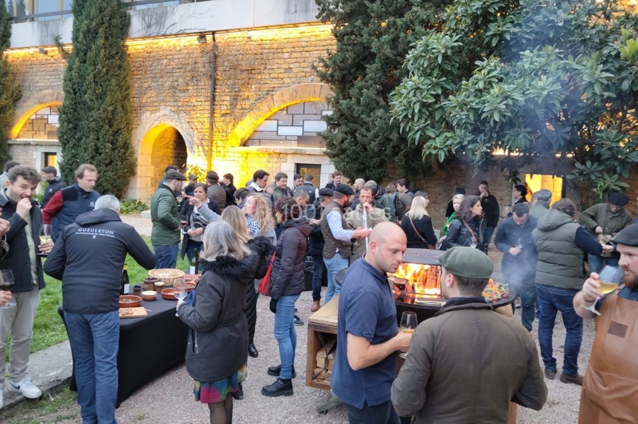 Groupe de personnes réunies en extérieur autour d'un buffet et d'un barbecue, dans une cour avec des murs en pierre éclairés.