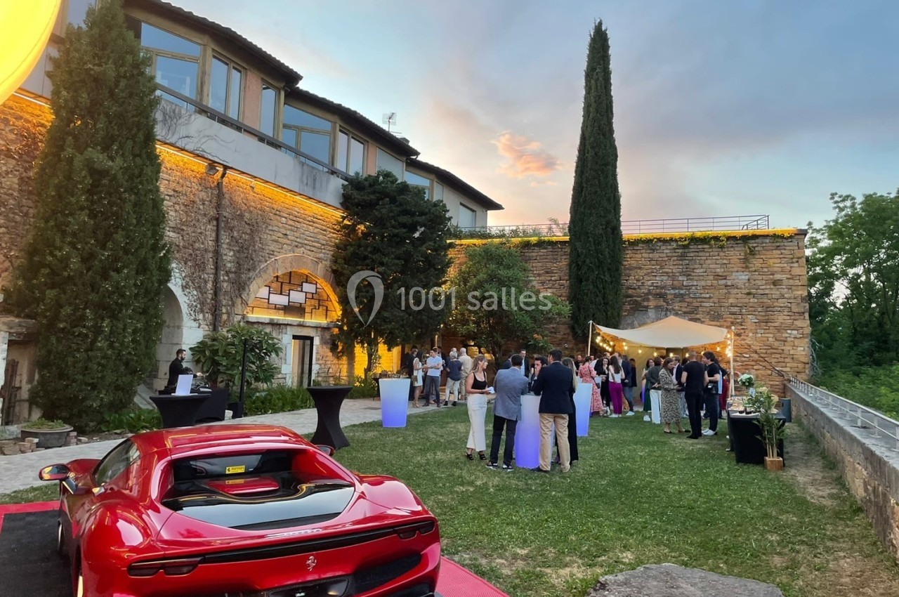 Groupe de personnes rassemblées dans un jardin près d'un bâtiment en pierre, avec une voiture rouge au premier plan.
