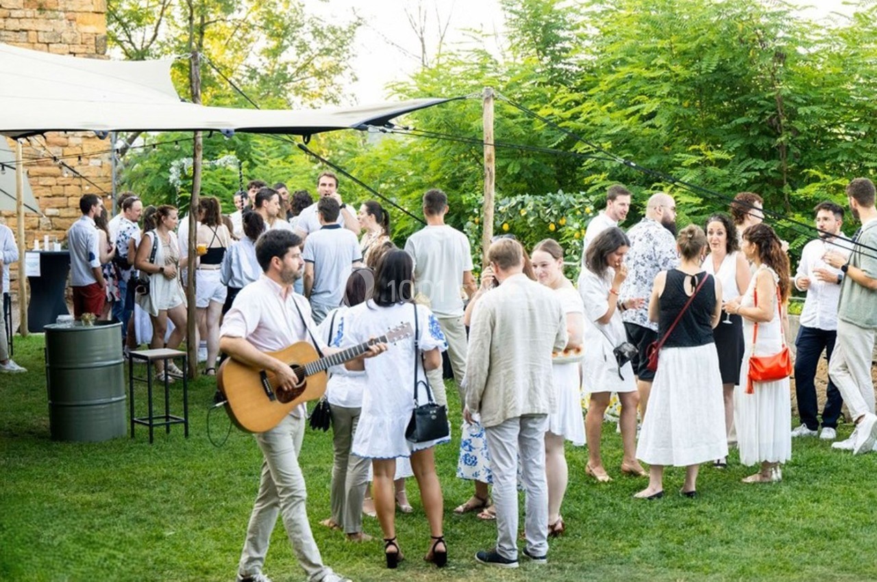 Groupe de personnes en tenue estivale discutant et écoutant un musicien jouant de la guitare dans un jardin verdoyant.