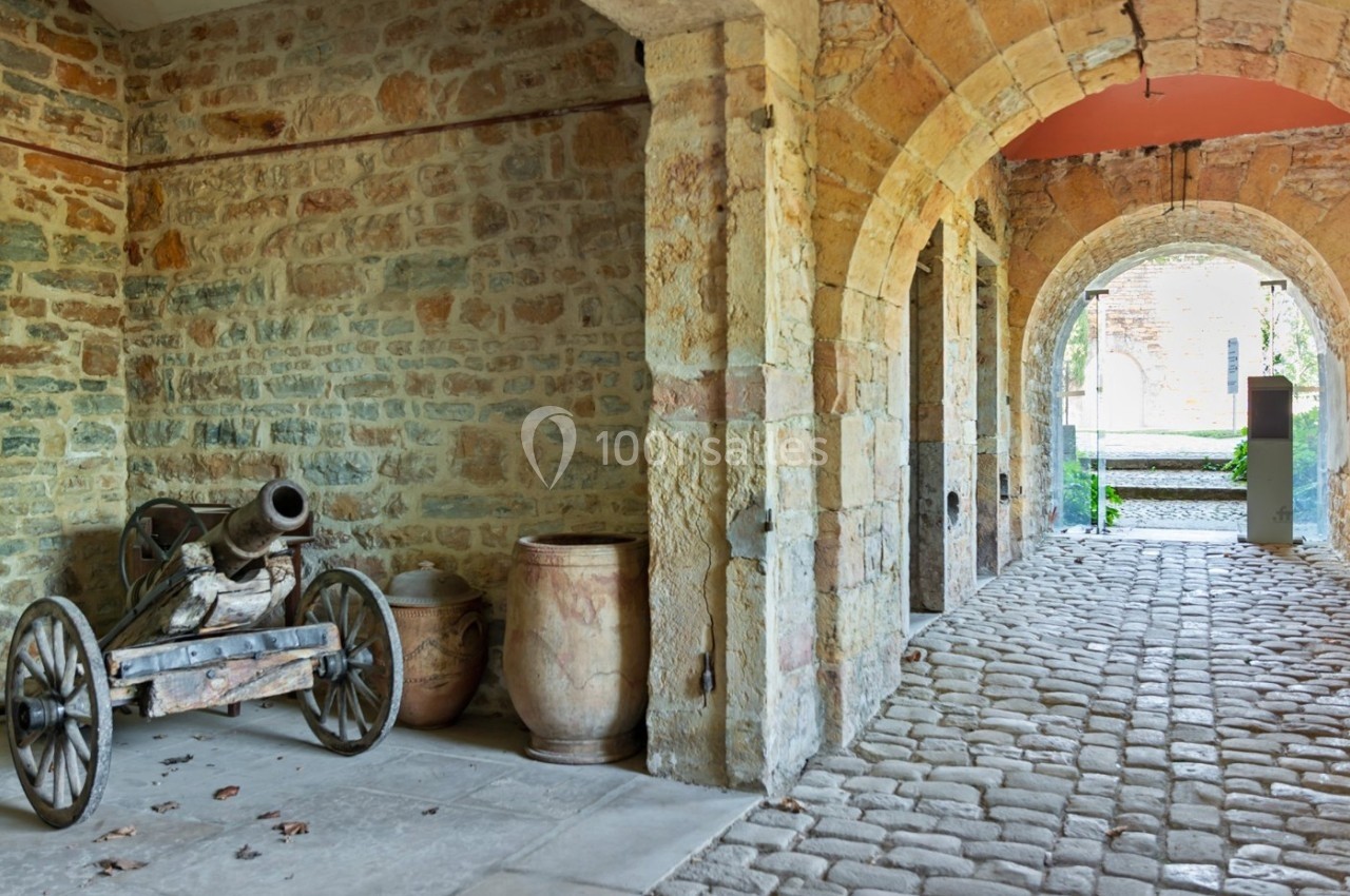 Un vieux canon sur roues et des jarres en terre cuite sont disposés sous une arcade en pierre dans une cour pavée.