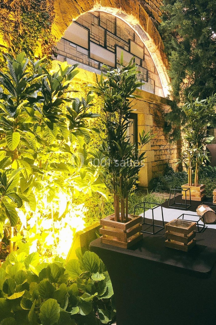 Espace extérieur aménagé avec des plantes, des tables noires et un éclairage chaleureux près d'une arche en pierre.