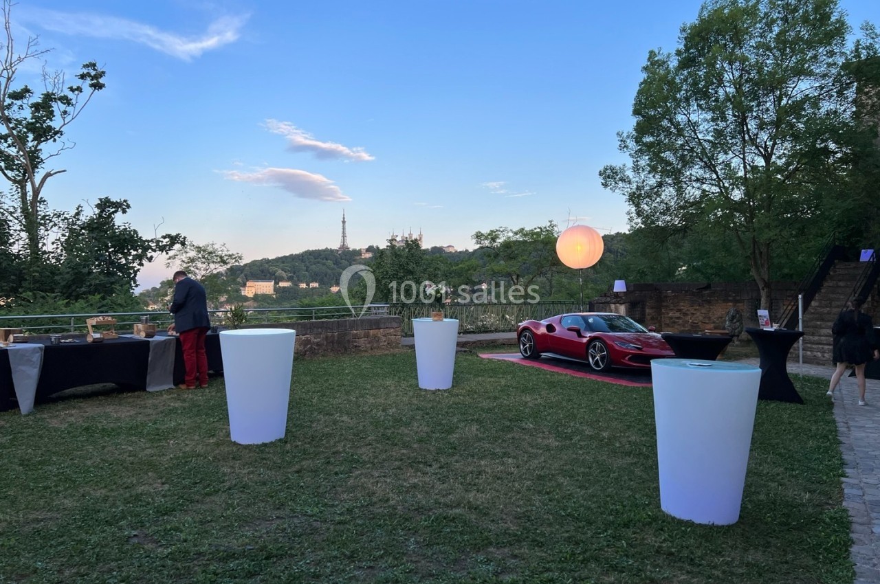 Vue d'un jardin avec une voiture sportive rouge, des tables hautes et un paysage de colline en arrière-plan au crépuscule.
