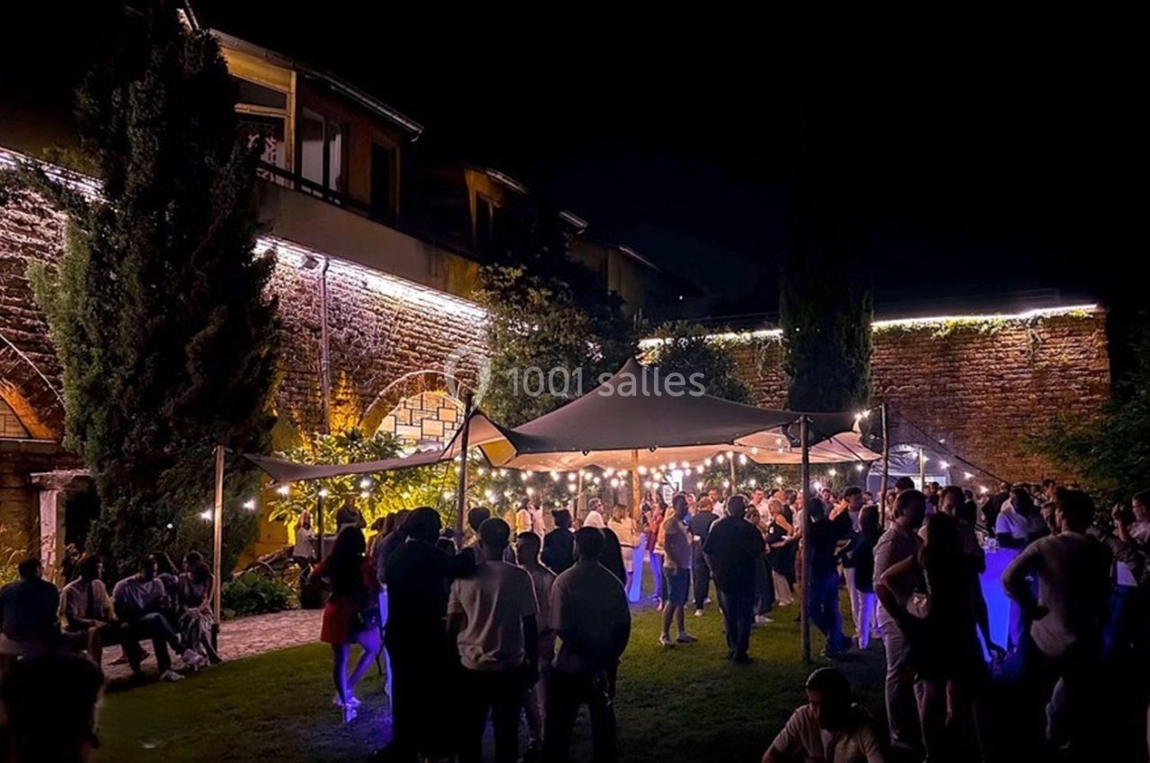 Groupe de personnes rassemblées sous un chapiteau éclairé dans un jardin, de nuit, avec des bâtiments en arrière-plan.