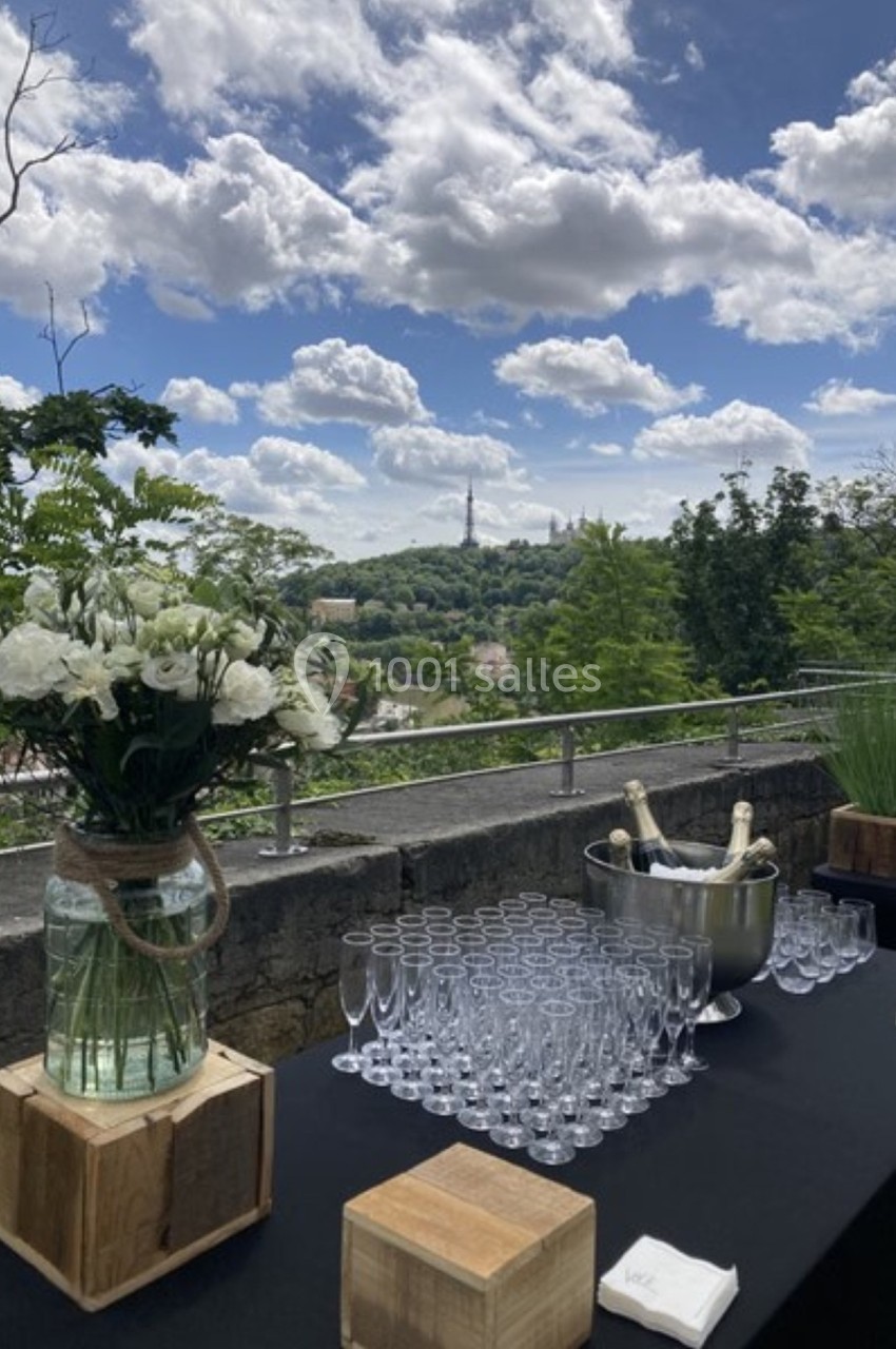 Table dressée en extérieur avec des fleurs, des verres à champagne et un seau à glace, vue sur une colline verdoyante.