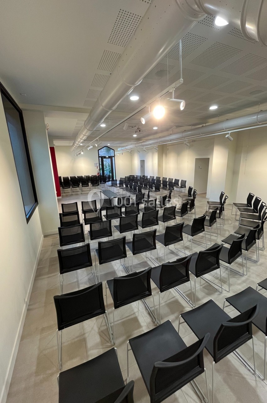 Salle de conférence lumineuse avec des rangées de chaises noires alignées face à une estrade.