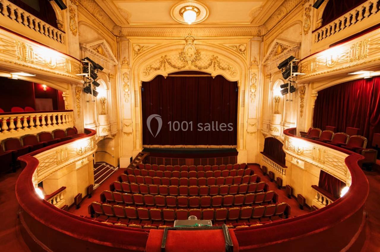 Intérieur d'un théâtre avec une scène fermée par un rideau rouge, entourée de balcons et de sièges vides.