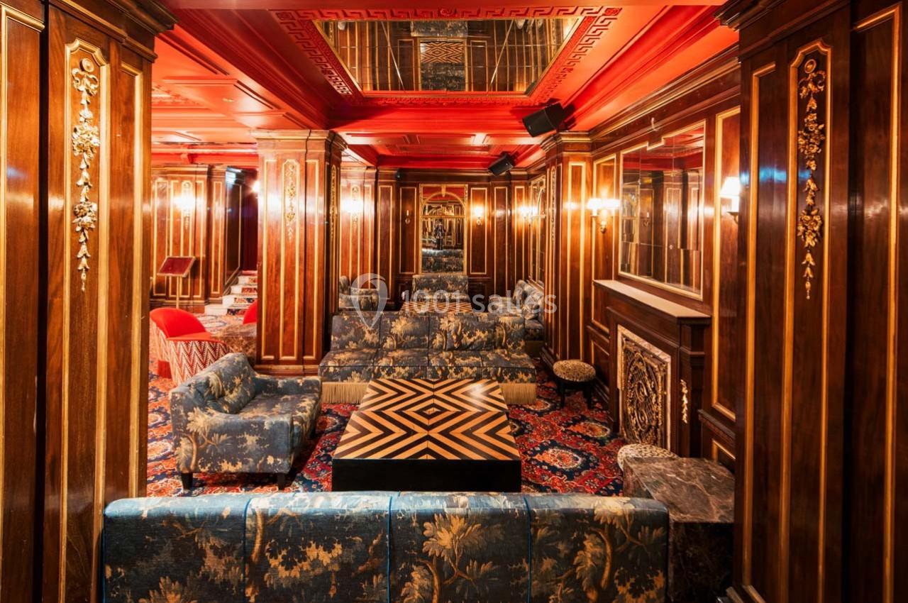 Salon élégant avec boiseries sculptées, canapés fleuris, tapis rouge à motifs et éclairage chaleureux.