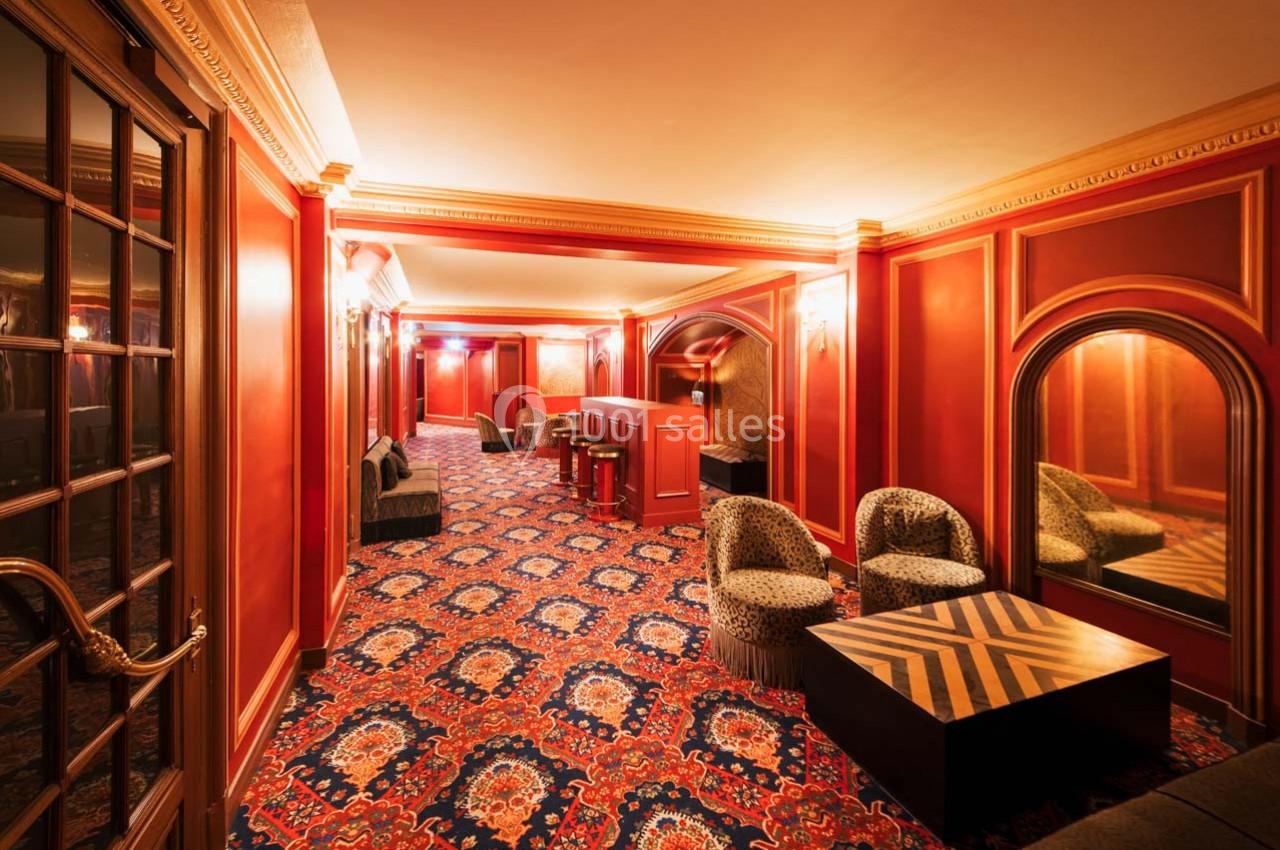 Salon élégant avec murs rouges, tapis à motifs, fauteuils, tables basses et miroirs dans un cadre chaleureux.