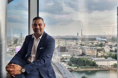 Homme souriant portant un casque audio et un costume sombre, éclairé par une lumière douce sur fond noir.