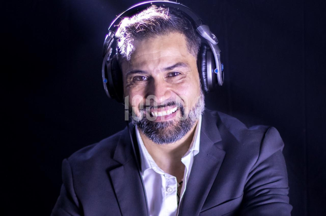 Homme souriant portant un casque audio et un costume sombre, éclairé par une lumière douce sur fond noir.