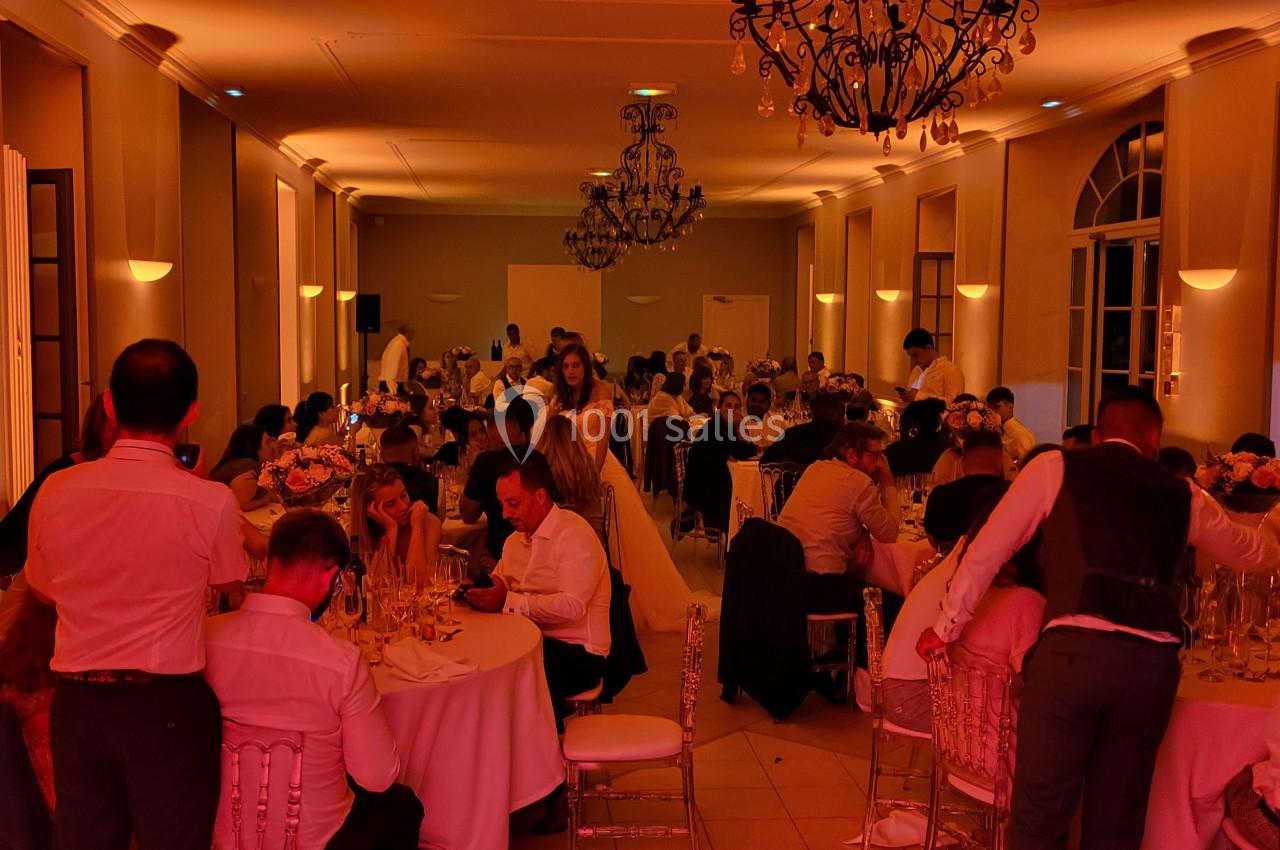 Salle de réception éclairée en rouge, avec des convives assis à des tables rondes lors d'un événement festif.