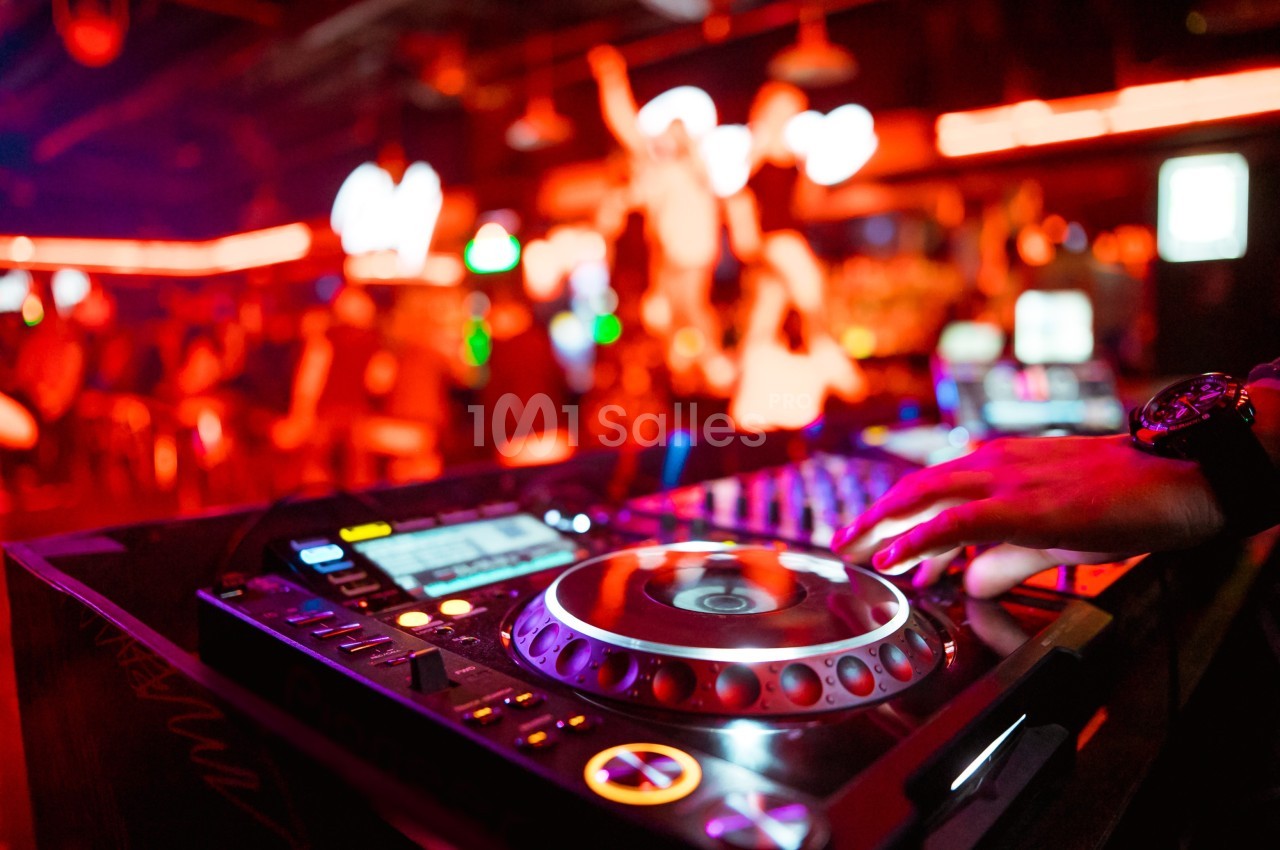 Main posée sur une table de mixage dans un club éclairé par des lumières rouges, avec une foule floue en arrière-plan.