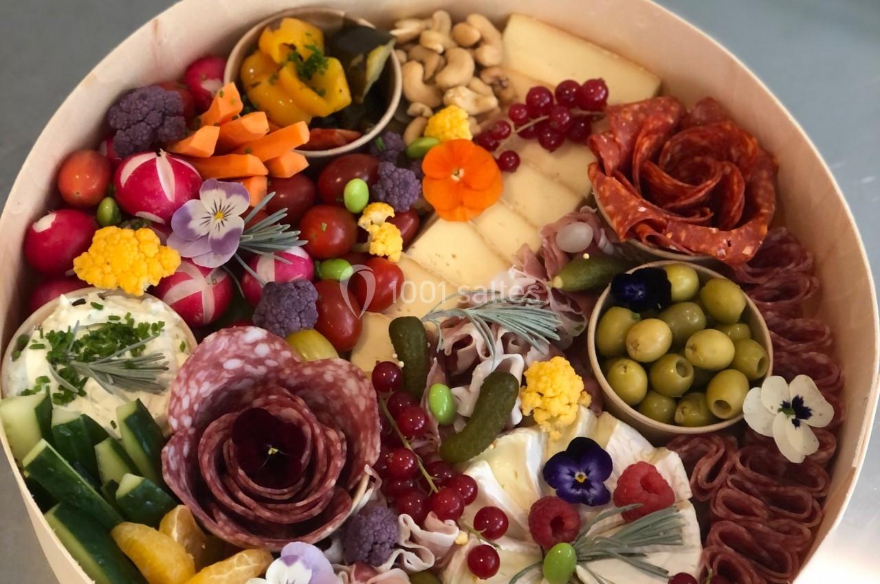 Plateau rond garni de charcuteries, fromages, fruits, légumes, olives et fleurs comestibles, disposés harmonieusement.