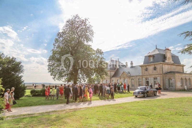 Un groupe de personnes rassemblées devant un château entouré de pelouses, avec une voiture ancienne garée à proximité.