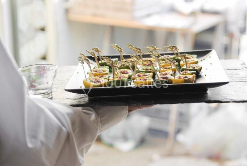 Plateau de bouchées apéritives présentées sur des cuillères en bois, porté par une personne en chemise blanche.