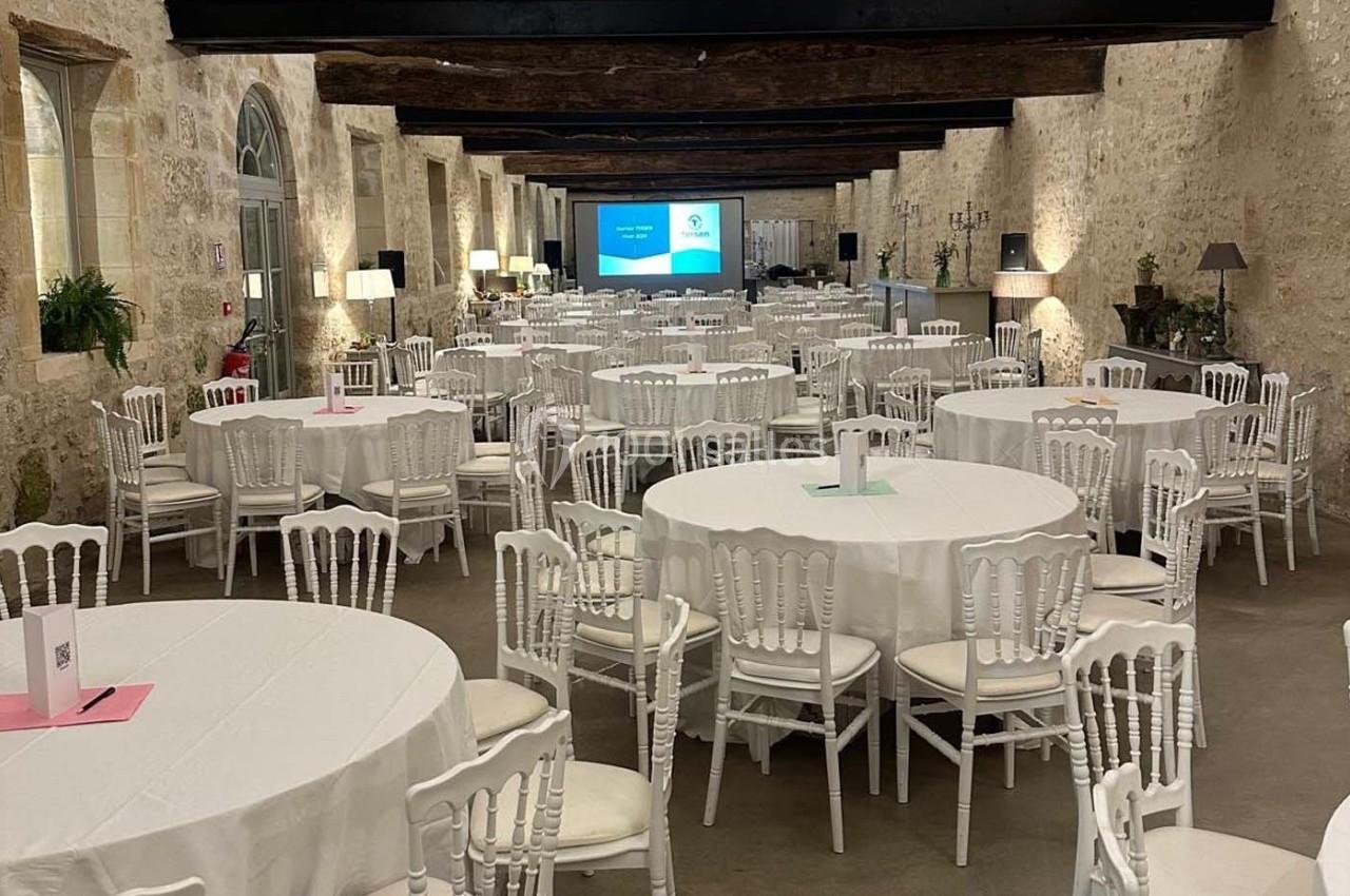 Salle de réception avec tables rondes dressées, chaises blanches et écran de projection au fond.