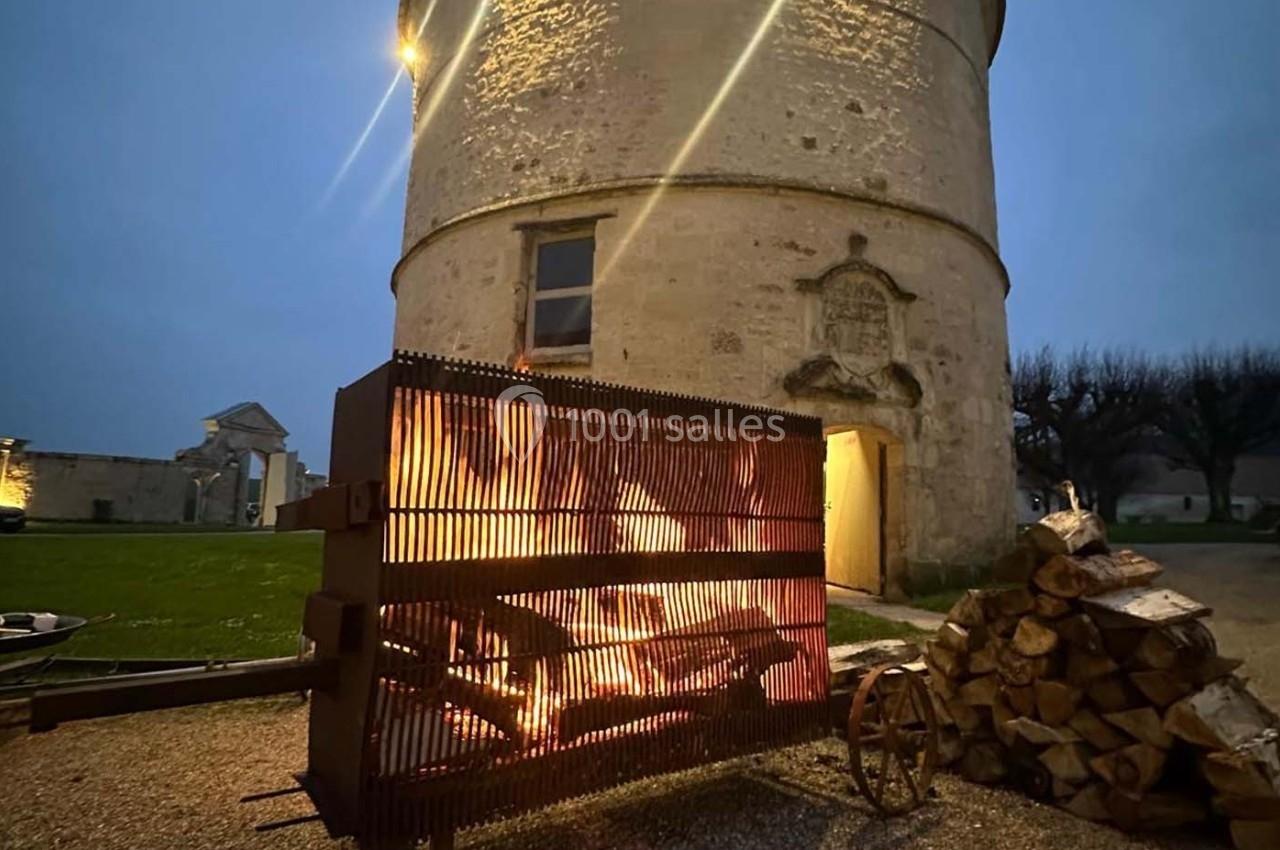 Brasero allumé devant une tour en pierre éclairée, avec des bûches empilées à proximité et un ciel crépusculaire.