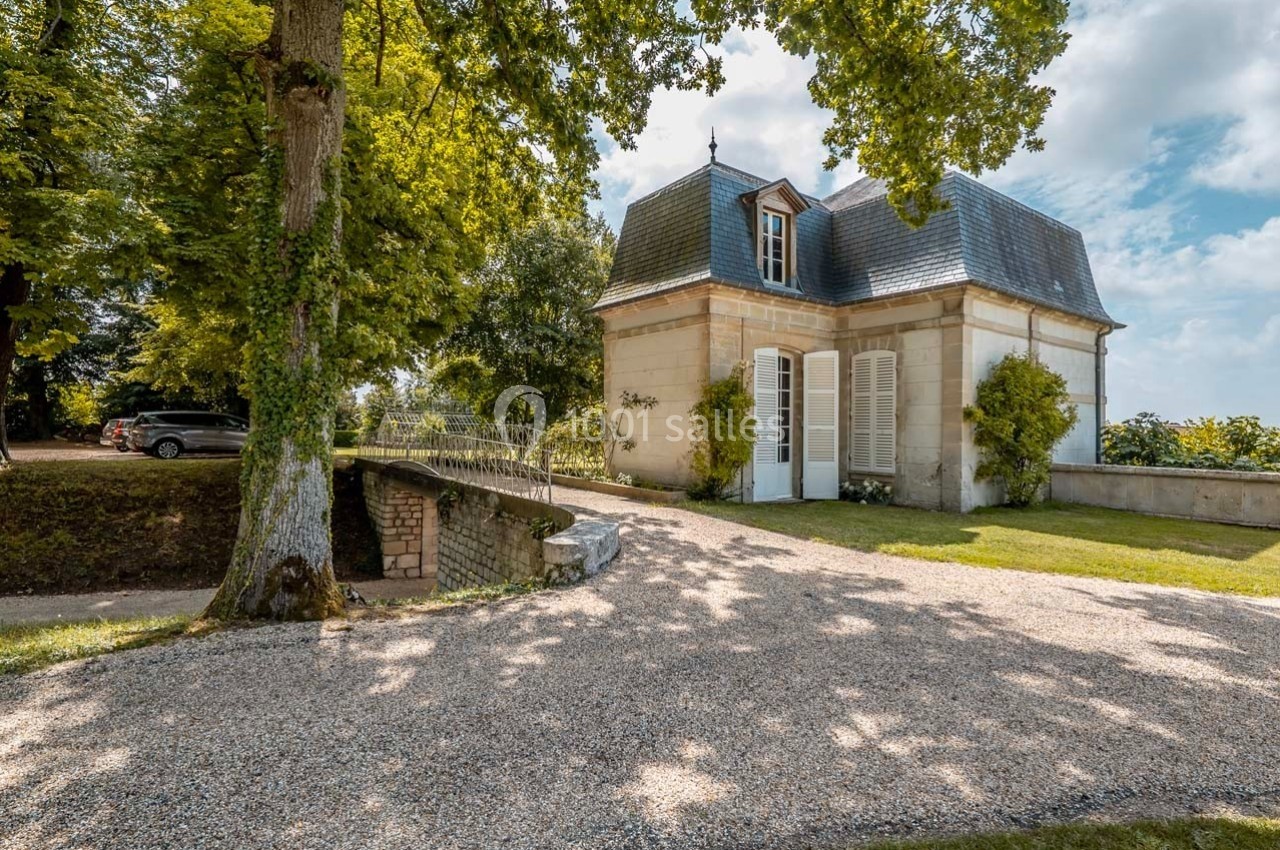 Petite maison en pierre avec volets blancs, entourée d'arbres et d'une allée gravillonnée, sous un ciel dégagé.