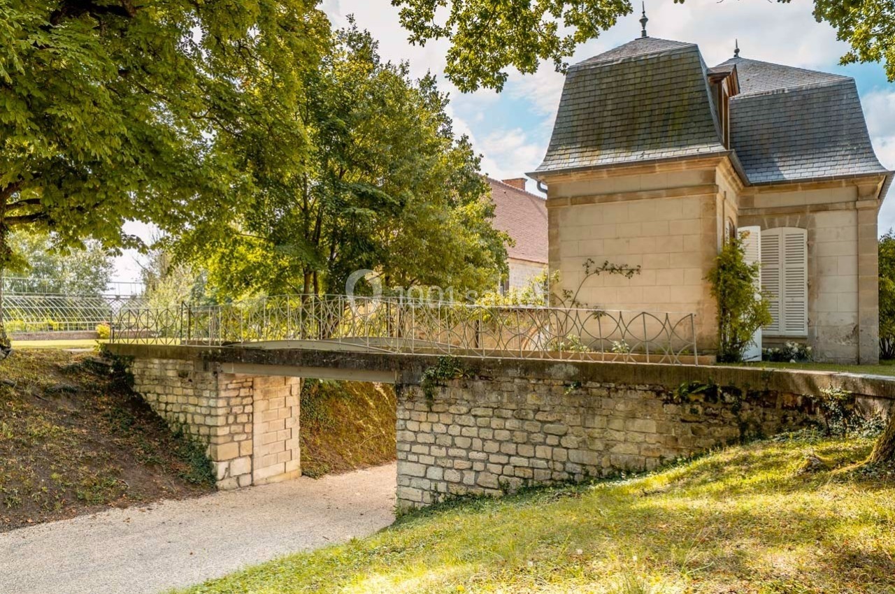 Petit pont en pierre et métal traversant un chemin, bordé d'arbres et menant à un bâtiment ancien avec toit mansardé.