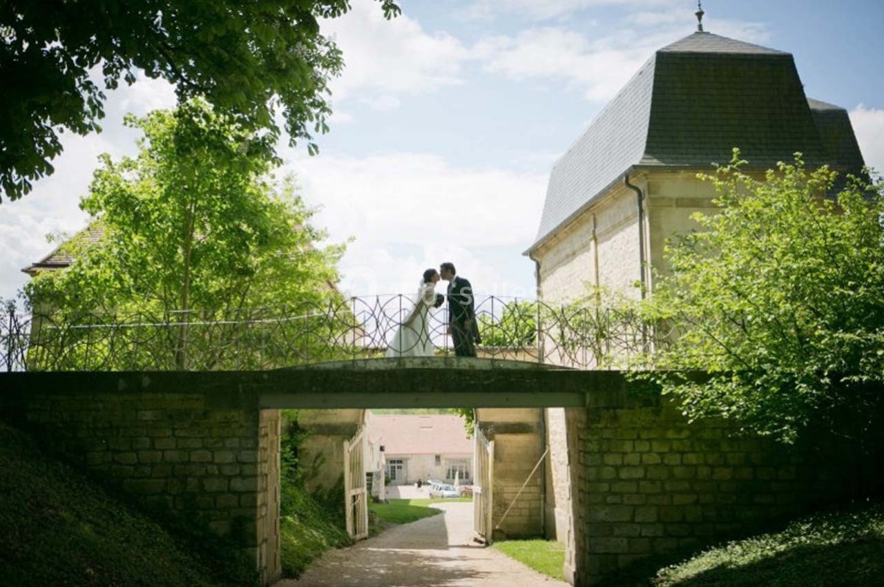 Un couple en tenue de mariage se tient sur un pont en pierre entouré de verdure, sous un ciel partiellement nuageux.