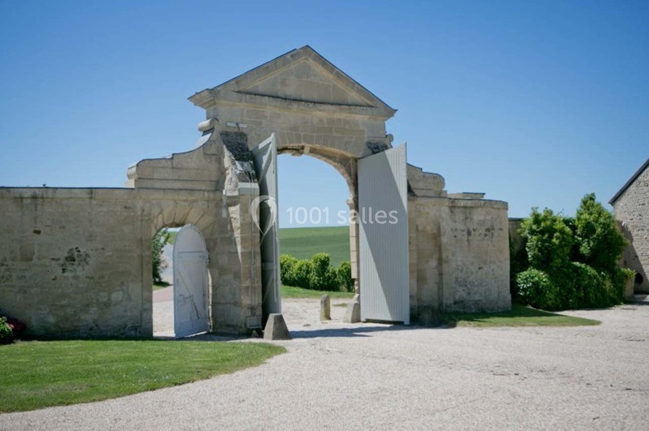 Portail en pierre ouvert sur une cour, entouré de murs et de verdure, sous un ciel bleu dégagé.