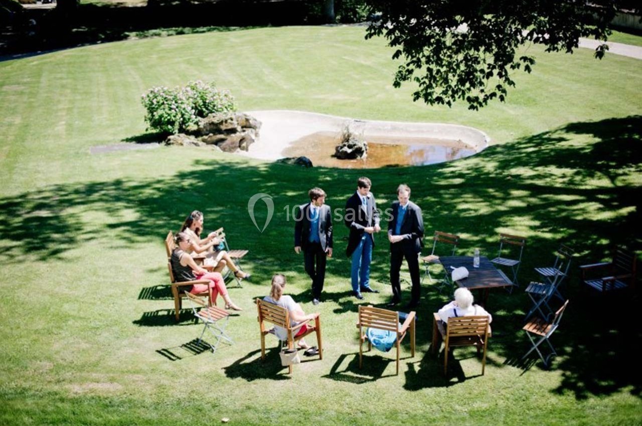 Des personnes discutent ou sont assises sur des chaises dans un jardin verdoyant avec un petit bassin en arrière-plan.