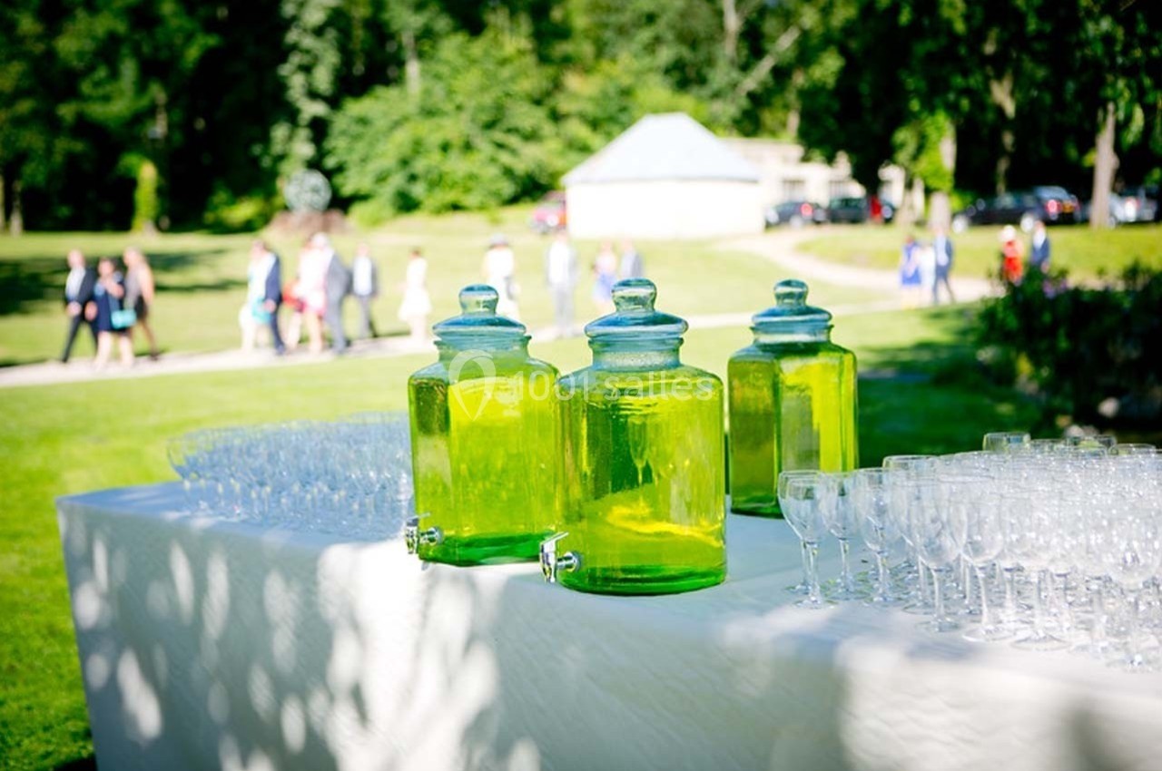 Trois bonbonnes de boisson verte sur une table blanche, entourées de verres, dans un parc ensoleillé.