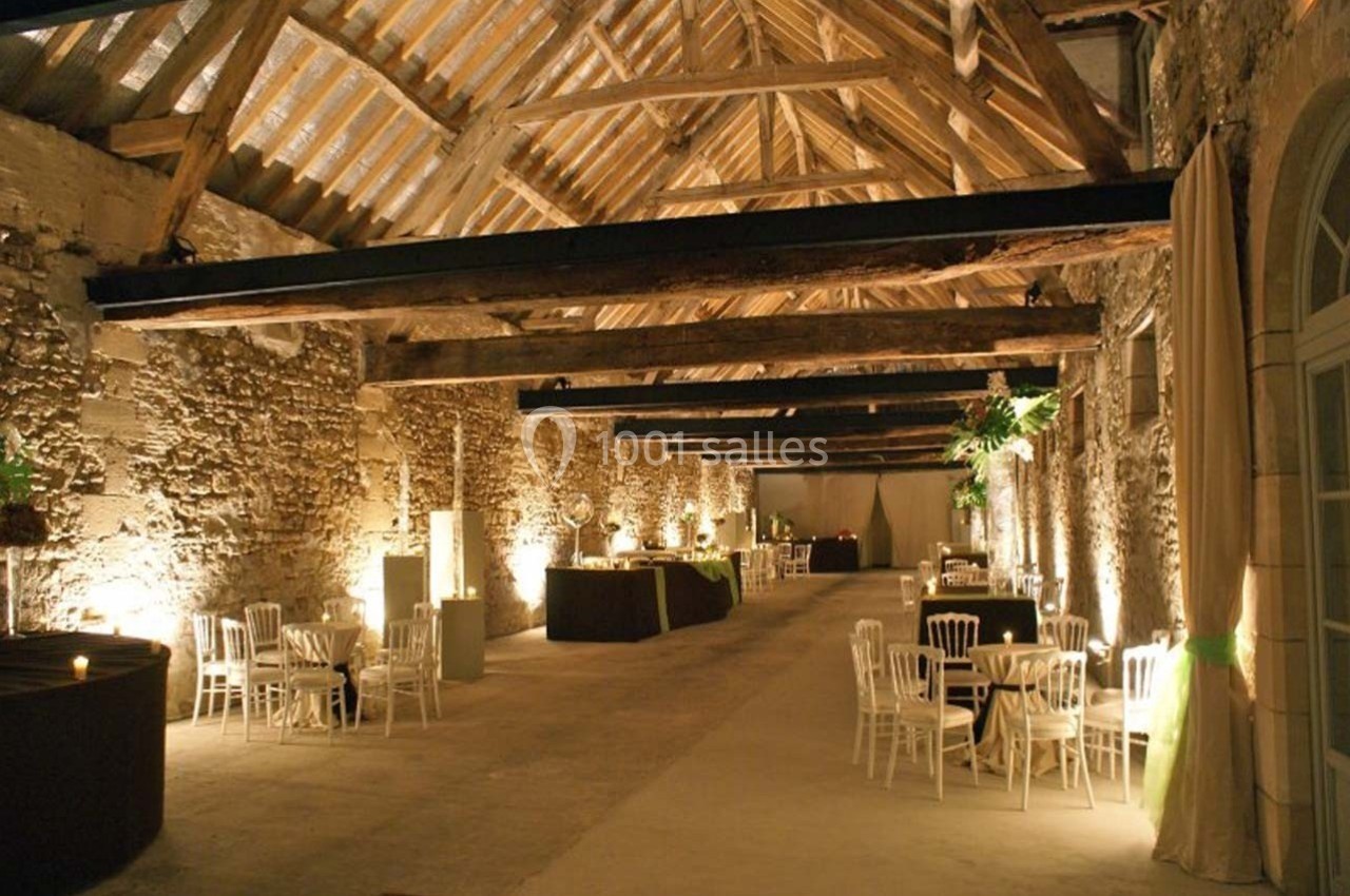 Salle en pierre avec poutres apparentes, aménagée pour un événement avec tables rondes et chaises blanches.